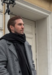 Szary płaszcz z wełnianej tkaniny o fakturze oraz czarny szalik. W tle zamknięte drzwi. Włosy lekko rozczochrane. Neutralne oświetlenie na zewnątrz.