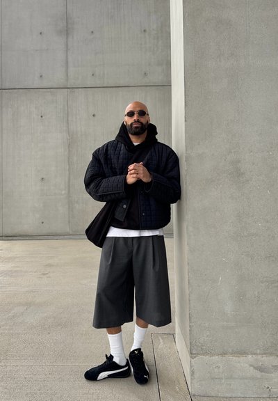 Chaqueta negra acolchada, superpuesta sobre una sudadera negra con capucha, combinada con unos pantalones cortos gris con pliegues, calcetines blancos y zapatillas negras. Fondo de hormigón liso.