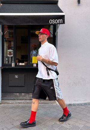 Persona caminando con una camisa blanca y pantalones cortos negros con rayas grises, calcetas rojas, zapatos negros y una gorra roja. Tienda negra de fondo.