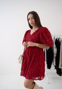Czerwona sukienka z nałożonymi rękawami i kwiatową fakturą, z dekoltem w kształcie litery V. Podkreślona małą beżow�ą torebką i białymi botkami.