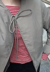 Graue, leichte Außenjacke mit Bindeband, die über einem rot-weißen gestreiften Hemd getragen wird. Schwarze Hosen sind am unteren Rand teilweise sichtbar.