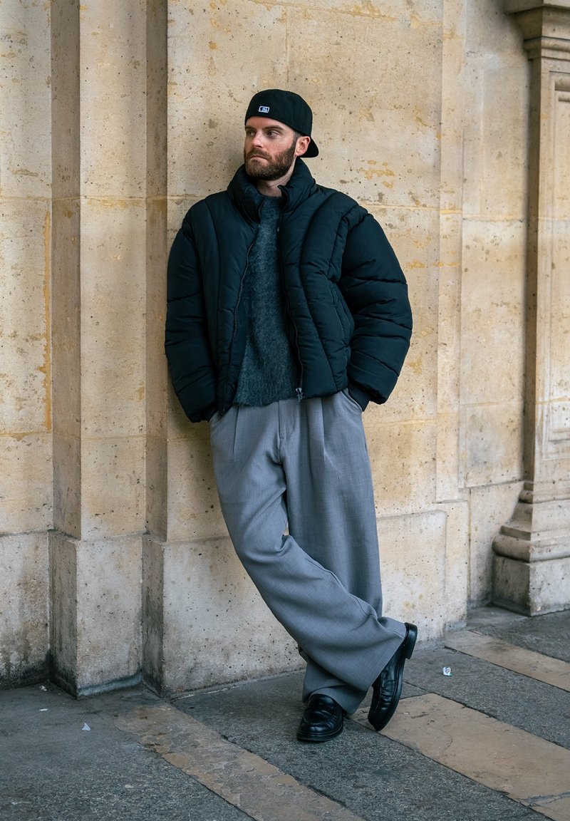 Homme portant une doudoune noire, un pantalon large gris, une casquette noire et des chaussures noires, appuyé contre un mur en pierre beige, les mains dans les poches.