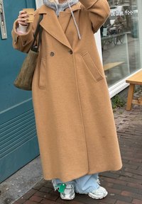 Personne en long manteau camel tenant une tasse de café, portant un sweat à capuche gris, un jean bleu clair et des baskets, debout près de l'entrée d'un café.