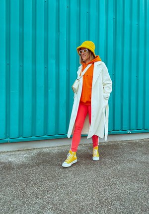Mujer con sombrero amarillo, abrigo blanco, sudadera con capucha naranja, pantalones rojos y zapatillas amarillas caminando frente a una pared de metal corrugado de color verde azulado.