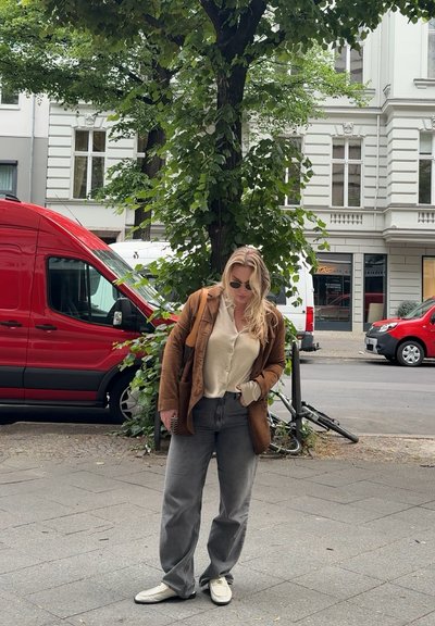 Chaqueta de cuero marrón, camisa beige de botones, pantalones grises holgados y zapatos blancos. De pie al lado de un árbol con una furgoneta roja de fondo.