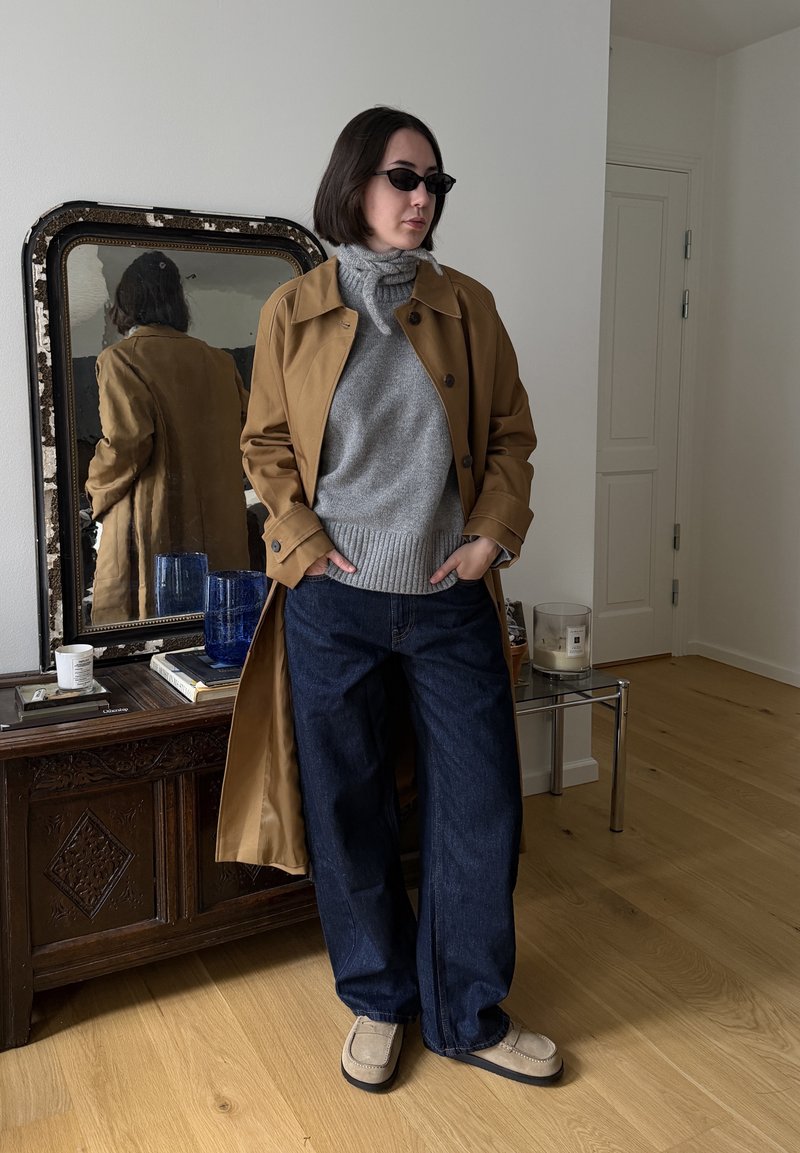 Personne portant des lunettes de soleil, un trench-coat beige, un pull à col roulé gris, un jean large foncé, debout les mains dans les poches dans une pièce avec un miroir et des meubles en bois.