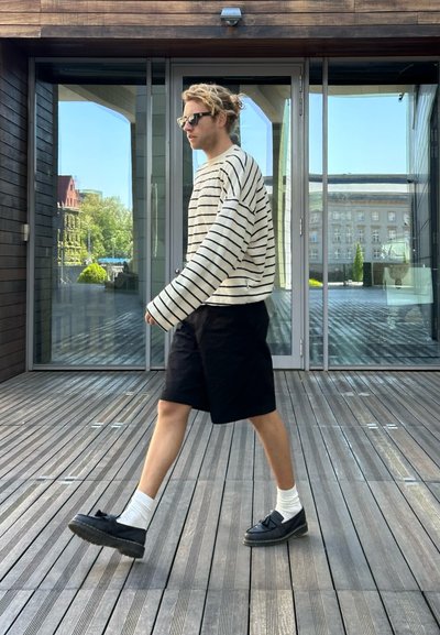 Suéter de rayas de manga larga en blanco y azul marino, combinado con pantalones cortos negros y zapatos oscuros. Terraza exterior de madera con grandes ventanas de vidrio.