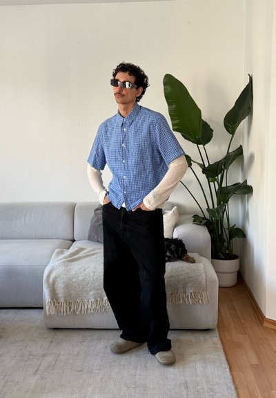 Hombre con gafas de sol, camisa de manga corta a cuadros azul sobre mangas largas blancas, pantalones negros, de pie en la sala junto a un sofá gris y una planta en maceta.