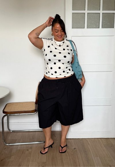 Mujer de pie en el interior, vistiendo una camisa blanca con lunares, pantalones cortos negros de pierna ancha, sandalias negras con tacón y llevando un bolso de hombro azul.