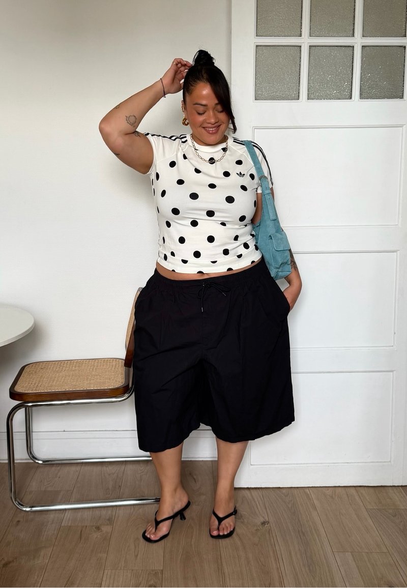 Femme debout à l'intérieur, portant une chemise blanche à pois, un short noir à jambes larges, des sandales à talons noires et portant un sac à épaule bleu.