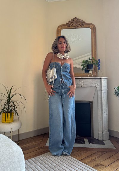 Mono de denim con diseño sin tirantes, color azul claro, frente abotonado y cintura con cordón. Bolso plateado, auriculares blancos y fondo de espejo.