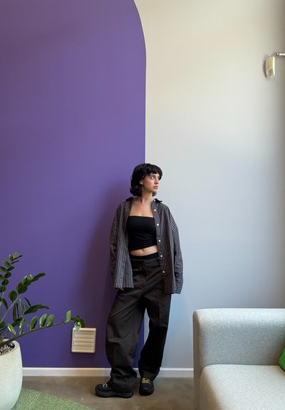 Camisa de rayas abotonada sobre un top corto negro, combinada con pantalones negros holgados y zapatos negros gruesos, frente a una pared morada y blanca.
