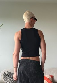 Crni rebrasti kratki top uparen s tamnim teksturiranim hlačama. Osoba ima kratku platinastu kosu i crne sunčane naočale. Minimalistička pozadina.