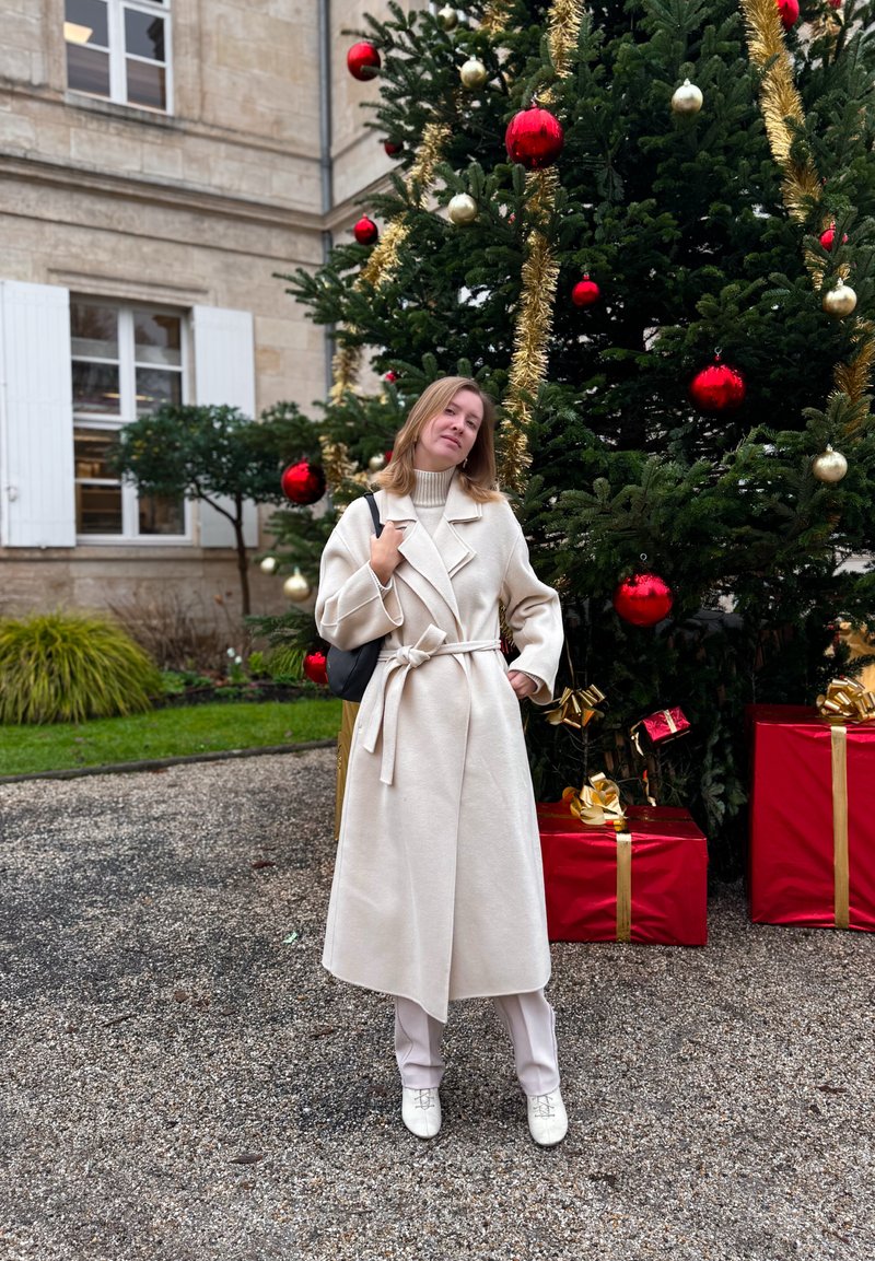 Manteau en laine beige avec larges revers, ceinture à la taille, associé à un pantalon crème et des chaussures blanches. Décorations de vacances en arrière-plan.