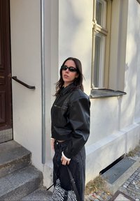 Donna in giacca di pelle nera e occhiali da sole, che tiene una borsa a fantasia zebrate, in piedi su gradini di pietra accanto a un edificio color crema con una finestra.