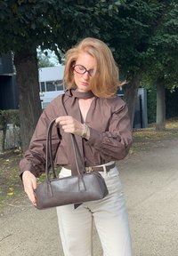 Ruskea nahkakäsilaukku, jossa on pitkä muoto ja tuplakahvat, esiteltynä sileällä pinnalla. Mallilla on päällään ruskea paita ja kermaväriset housut.