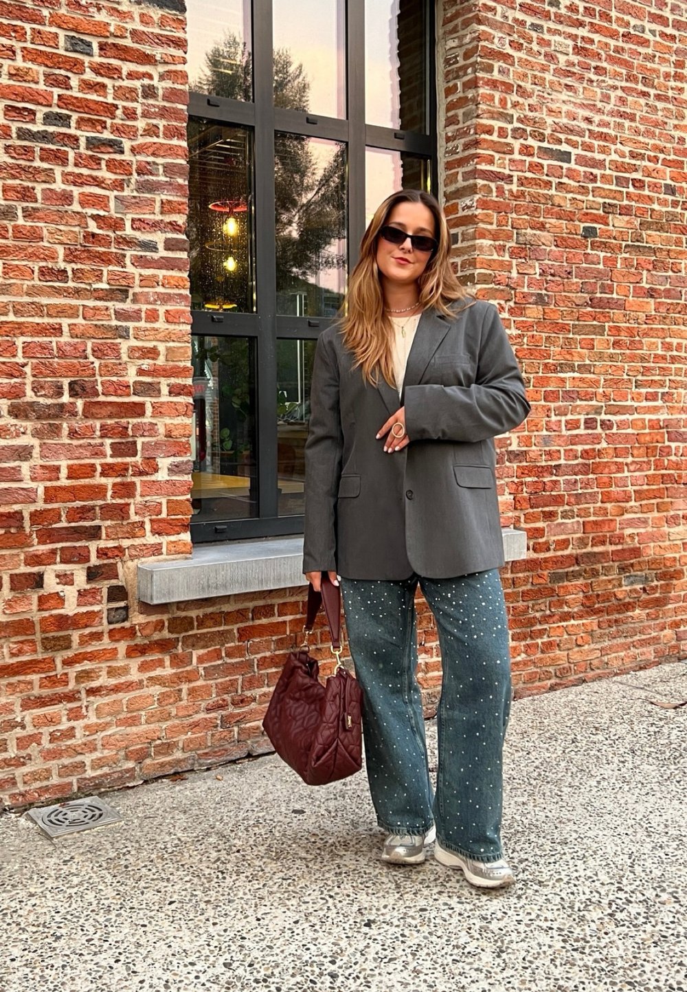 Γκρι υπερμεγέθη σακάκι, κρεμ τοπ, διακοσμημένο τζιν σε μπλε απόχρωση, μεταλλικά αθλητικά παπούτσια και μπορντό τσάντα μπροστά σε φόντ�ο από τούβλα.