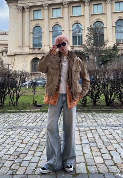 Joven con cabello rosa que lleva gafas de sol, chaqueta beige sobre camisa de cuadros naranja, camiseta blanca, vaqueros de pierna ancha y zapatillas negras, de pie sobre un camino de adoquines.