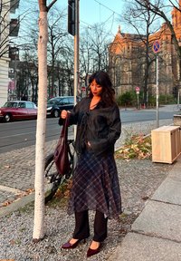 Svart teksturert jakke med snøring foran, lagd over et blått rutete skjørt. Sorte bukser og burgunderfargede spisse hæler fullfører antrekket.