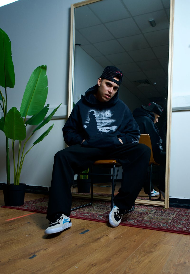 Schwarzer Hoodie mit grauem Aufdruck, oversized Schnitt, kombiniert mit schwarzen Hosen und mehrfarbigen Sneakers. Grüne Blätter und ein Spiegel im Hintergrund.