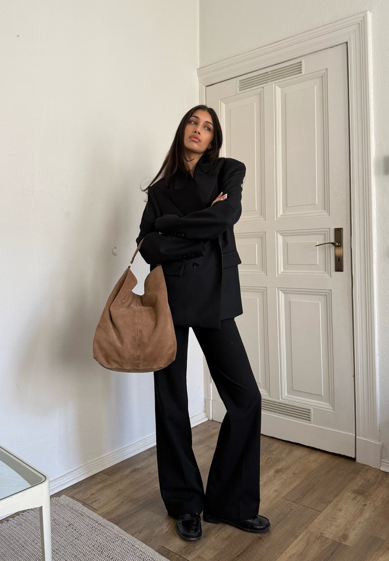 Γυναίκα με μαύρο κοστούμι και παπούτσια που κρατά μεγάλο καφέ σουέντ τσάντα ώμου, στέκεται μέσα σε κλειστό χώρο δίπλα σε λευκή πόρτα με πάνελ σε ξύλινο πάτωμα.