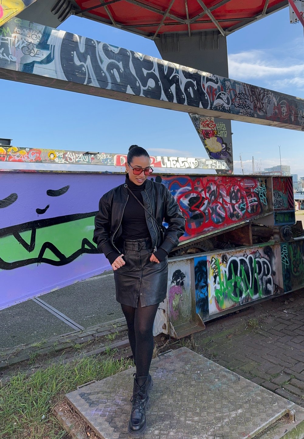 Čierna kožená bomber bunda cez čierny rolák, doplnená čiernou koženou sukňou. Šedé patterned platform topánky. Graffiti pozadie.