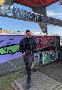 Čierna kožená bomber bunda cez čierny rolák, doplnená čiernou koženou sukňou. Šedé patterned platform topánky. Graffiti pozadie.