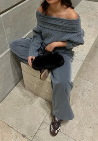 Personne assise sur un banc en pierre portant un pull gris épaules dénudées, un pantalon large gris, des baskets marron et blanches, tenant un petit sac à main noir en fourrure.