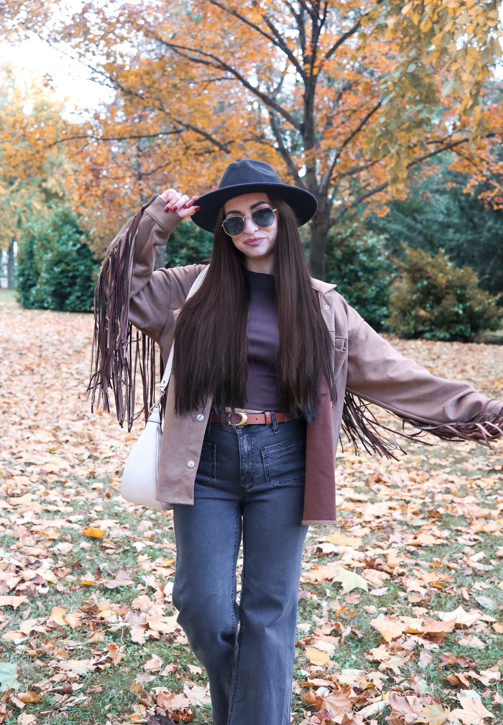 Bruine jas met franjes, donker topje, donkerblauwe high-waisted jeans, zwarte hoed met brede rand, ronde zonnebril; gefotografeerd tegen een achtergrond van herfstblad en bladeren.