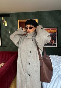 Ženský voľný šedý kabát s vysokým golierom a veľkými gombíkmi, doplnený čiernym klobúkom a hnedou koženou taškou. Priehľadné slnečné okuliare dotvárajú vzhľad.