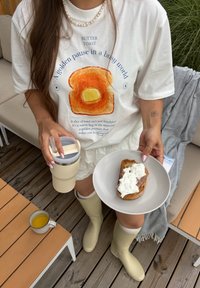 Hvit t-skjorte med et toast-motiv, som holder en beige kopp med lokk, og en tallerken med brød toppet med krem. Kombinert med beige støvler til kneet.