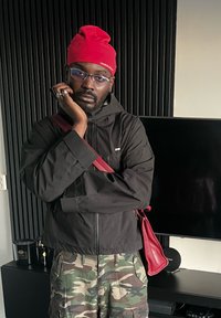 Veste noire zippée, shorts camouflage, bonnet rouge, lunettes, tenant le menton. Sac bandoulière rouge reposant sur l'épaule. Fond sombre avec des objets.