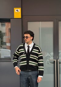 Cardigan rayé noir et vert avec des boutons sur le devant, chemise blanche à col, et cravate foncée. Lunettes de soleil noires et écouteurs blancs. Jean bleu.