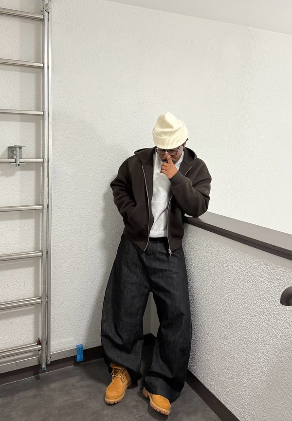 Person wearing white beanie, glasses, dark zip-up hoodie, wide black pants, and tan boots, leaning against white wall by metal ladder.