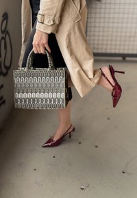 Rote Lackleder Slingbacks mit spitzen Zehen und dünnem Absatz; graue, gemusterte Handtasche mit gewölbten Designs, die von einer Hand gehalten wird.