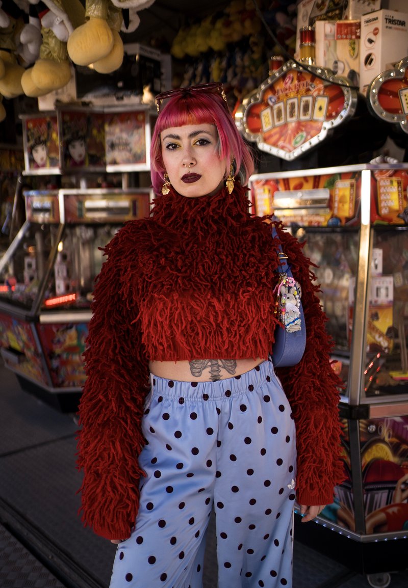 Personne aux cheveux roses et au rouge à lèvres foncé portant un pull rouge duveteux et un pantalon bleu clair à pois, debout devant des machines d'arcade.