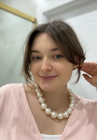 Collana di perle bianche con grandi perle lucide, indossata sopra un top rosa chiaro a costine. L'illuminazione soffusa e lo sfondo sfocato migliorano la messa a fuoco.