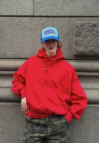 Sudadera roja de gran tamaño con un bolsillo canguro en el frente y cordón ajustable. Combinada con pantalones cargo de camuflaje y una gorra trucker azul.