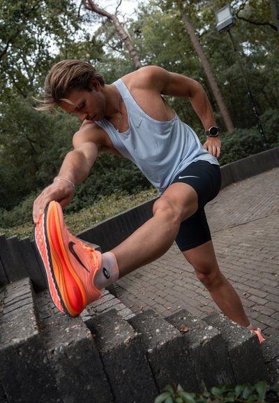Hombre atlético estirándose al aire libre, vistiendo una camiseta sin mangas de color azul claro y pantalones cortos negros, con zapatillas de correr de color naranja brillante. Entorno natural.