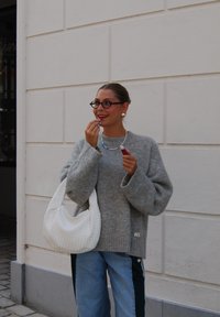 Suéter de punto gris, jeans azul con rayas laterales, bolso de mano tejido blanco, collar colorido, sosteniendo un lápiz labial, con gafas y pendientes.