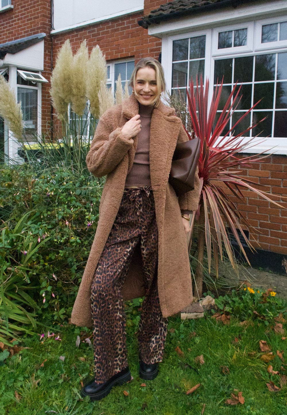 Brown teddy coat over a brown fitted top and leopard-print wide-leg trousers. Black platform shoes, carrying a dark brown bag. Green plants in the background.