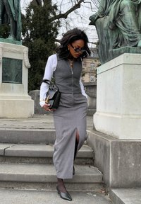 Grauer ärmelloser Weste mit Reißverschluss über einem weißen Hemd, kombiniert mit einem grauen, gestreiften Rock mit Schlitz vorne. Schwarze Handtasche und Schuhe.
