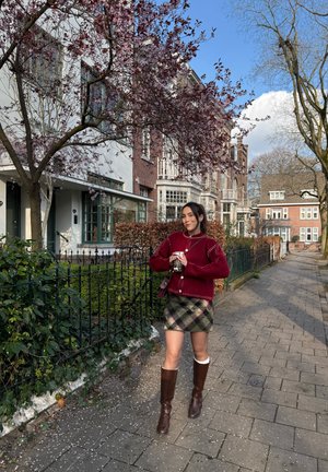 Mujer de pie en una acera en un barrio residencial sosteniendo una taza, vistiendo un suéter rojo, falda de cuadros y botas marrones.