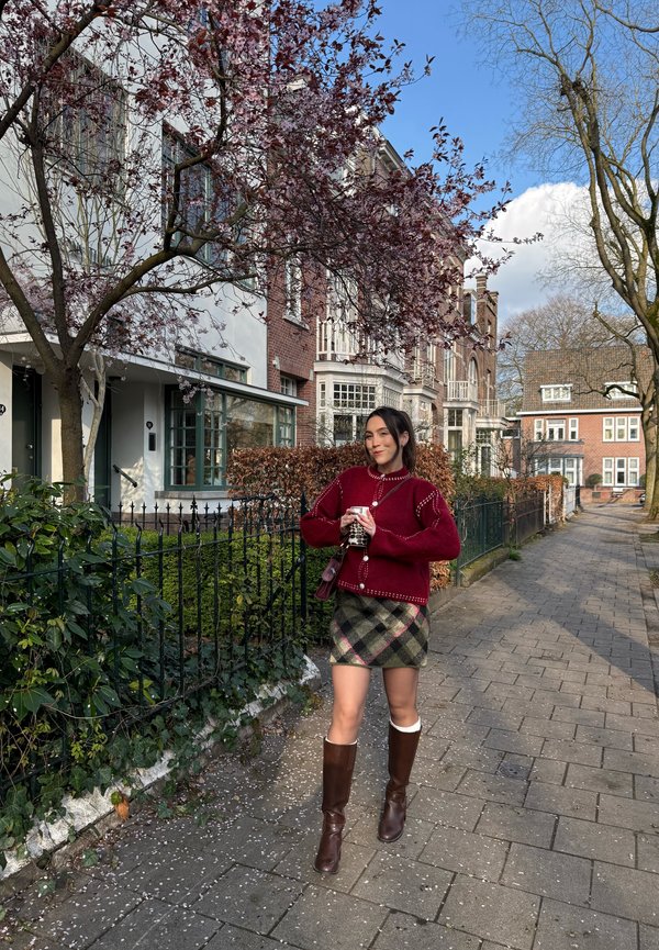 Kobieta stojąca na chodniku w dzielnicy mieszkalnej, trzymająca kubek, ubrana w czerwony sweter, kratowaną spódnicę i brązowe buty.