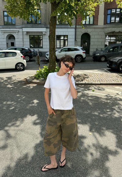 Una camiseta blanca corta, pantalones cortos cargo de camuflaje y chanclas negras. El conjunto presenta un ajuste relajado y un tejido liviano.
