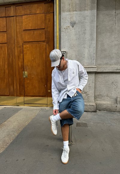Camisa de botones a rayas blancas, pantalones cortos de denim azul, zapatillas blancas y gorra. Cadena de plata notable y calcetines visibles. Entorno urbano.