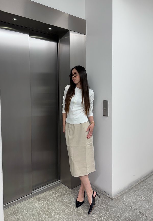 Mujer con cabello largo, gafas, top blanco, falda beige y tacones negros, de pie junto a un ascensor cerrado de acero inoxidable en el pasillo de un edificio moderno.
