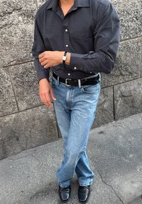 Camisa de manga larga gris oscuro, jeans de denim azul, cinturón negro, reloj plateado. Enmarcado contra una pared de piedra texturizada y pavimento.