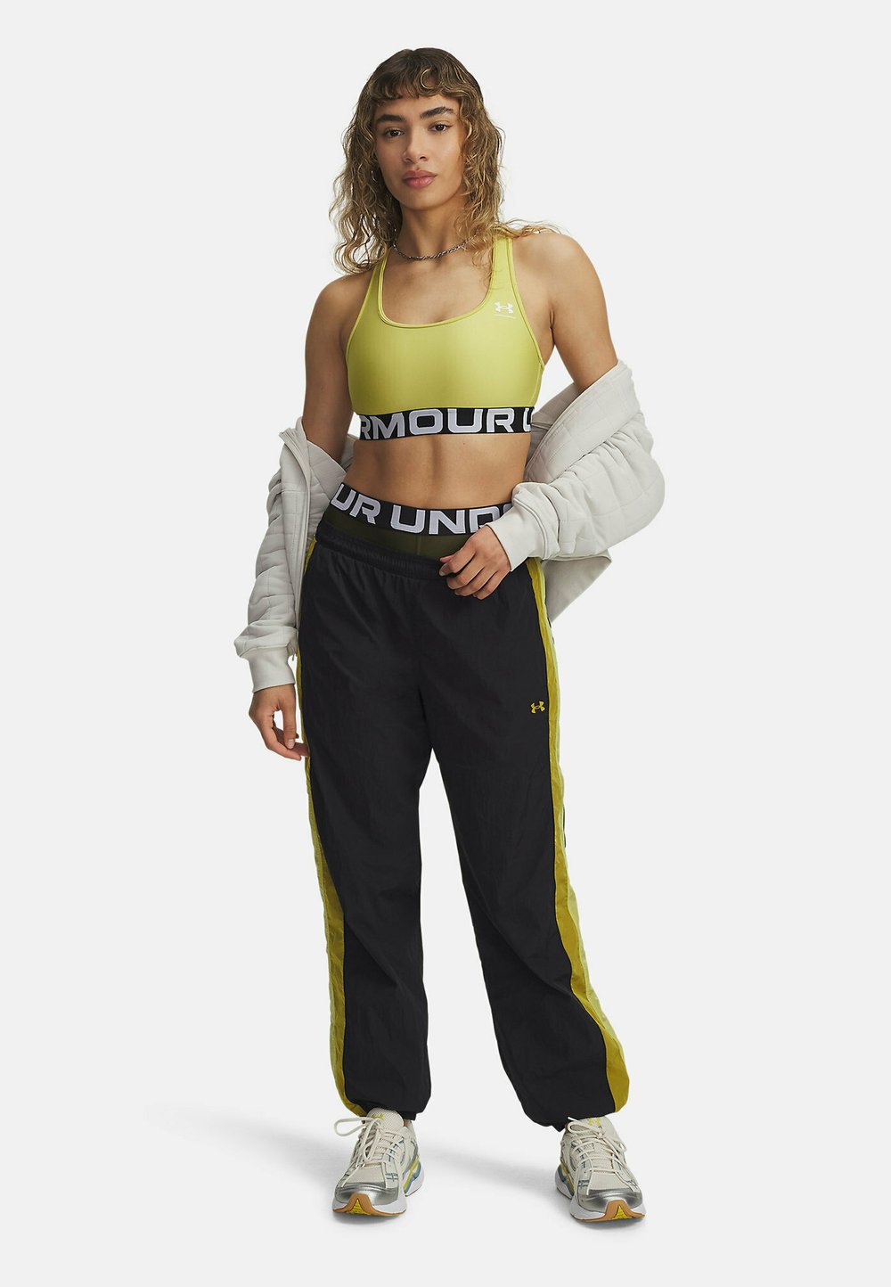 Žltá športová podprsenka s čiernym pásom, čierne nohavice na joging s žltými pruhmi po stranách, biela bunda prehodená cez ramená, šedé tenisky s akcentmi.