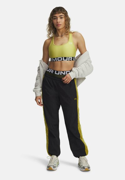 Sujetador deportivo amarillo con banda negra, pantalones joggers negros con rayas laterales amarillas, chaqueta blanca caída sobre los hombros, zapatillas grises con detalles.